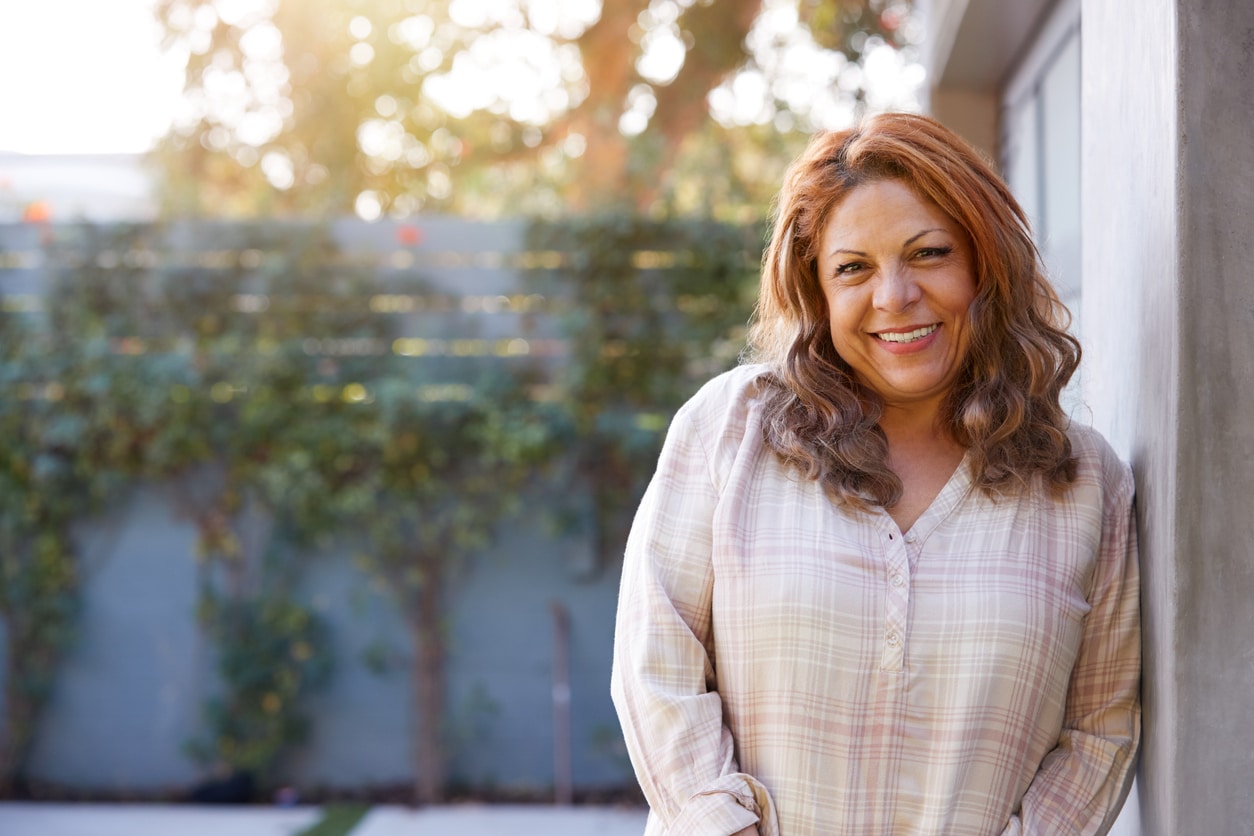 Portrait Of Smiling Senior Hispanic Woman In Garden At Home Against Flaring Sun